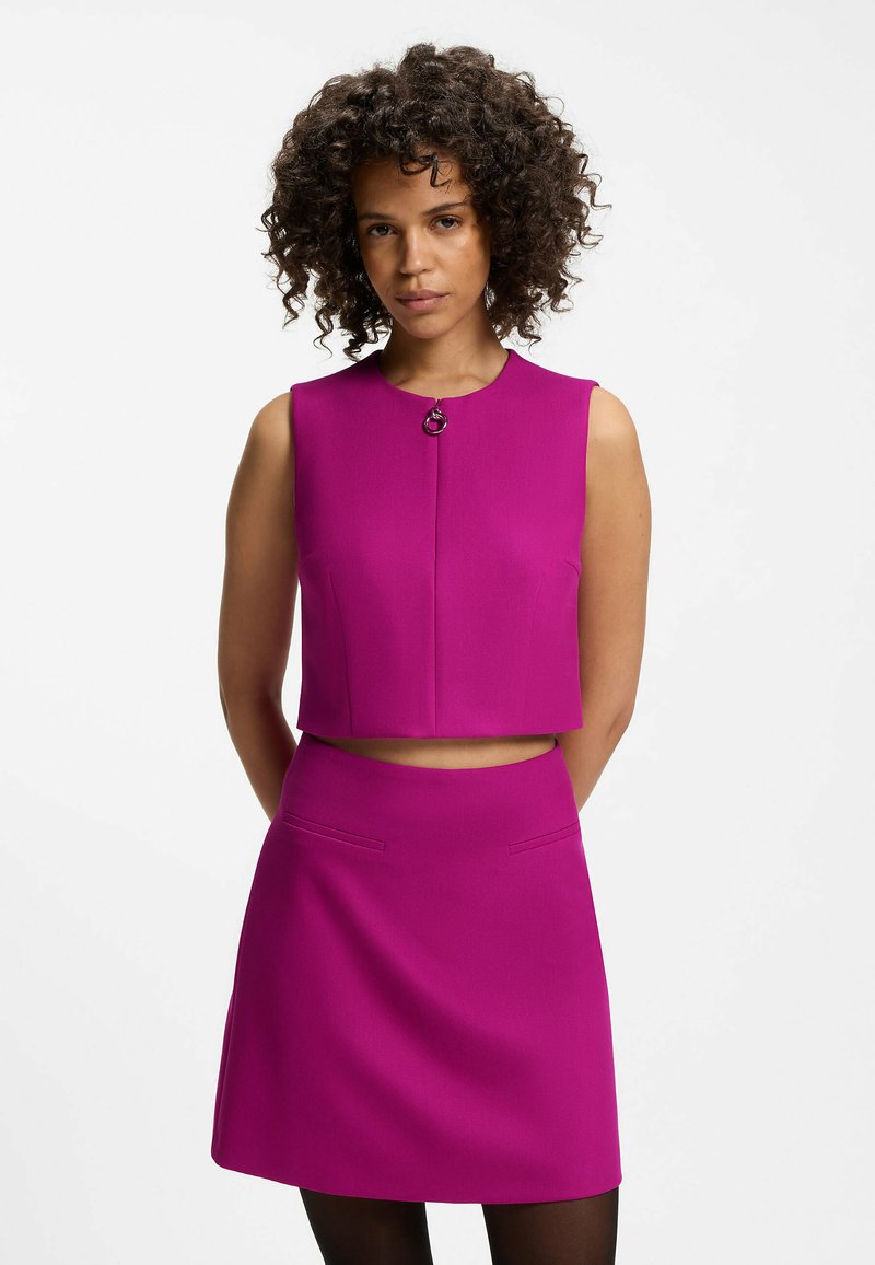 Femme aux cheveux bouclés portant un crop top sans manches magenta et une jupe assortie, debout, les mains dans le dos, sur un fond uni.