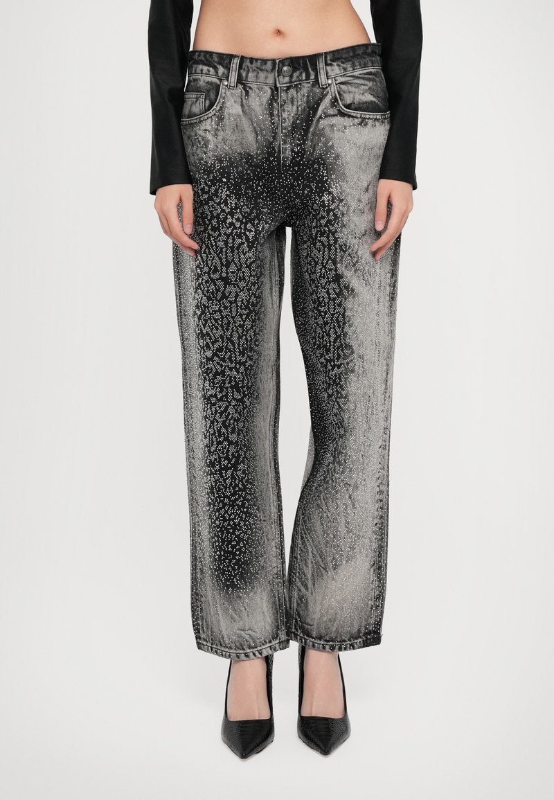 Woman wearing high-waisted wide-leg jeans with a black and silver metallic gradient and rhinestone pattern, paired with black pointed heels.