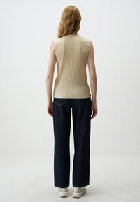 Top beige senza maniche con dettagli a righe verticali, abbinato a jeans a gamba larga blu scuro e sneaker bianche. Sfondo neutro.