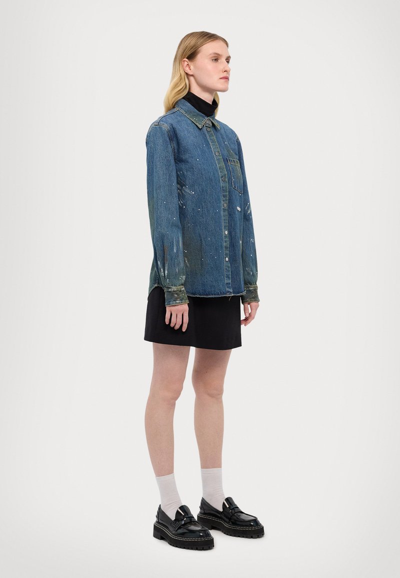 Cămașă din denim cu guler, având un buzunar frontal și detalii de stropi de vopsea. Asortată cu o fustă neagră și pantofi loafers negri cu tălpi groase.