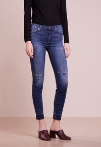 Jeans ajustados azules con detalles desgastados y un lavado medio, combinados con zuecos oscuros de tacón. Top negro de manga larga sencillo.