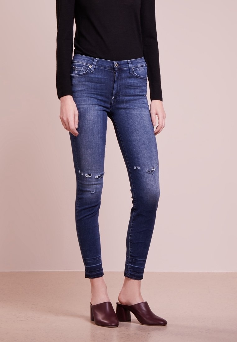 Jeans ajustados azules con detalles desgastados y un lavado medio, combinados con zuecos oscuros de tacón. Top negro de manga larga sencillo.