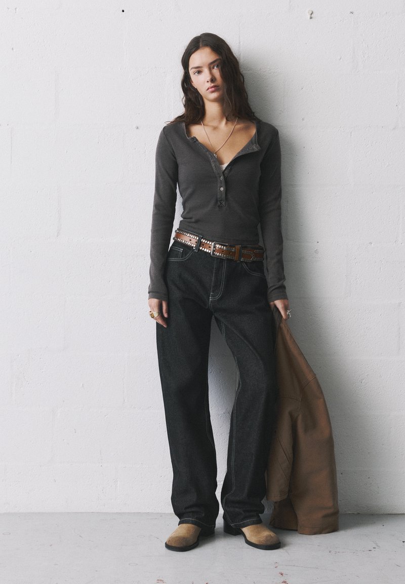 Langärmliges graues Jersey-Oberteil mit Knöpfen, schwarze weite Jeans, brauerN studierter Gürtel und beigen Slipper. Braune Jacke über dem Arm drapiert.