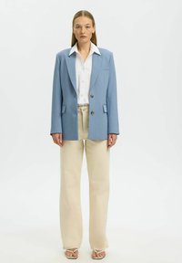 Femme debout de face portant un blazer bleu clair, une chemise blanche, un pantalon beige à jambes larges et des sandales blanches à bout ouvert sur un fond uni.