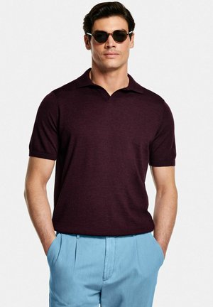 Uomo con polo a maniche corte viola scuro, pantaloni azzurri con le mani nelle tasche e occhiali da sole scuri.