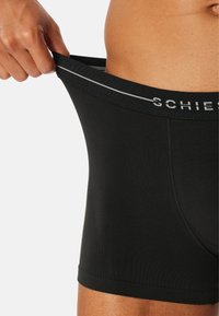 Mand iført sorte boxershorts, trækker i linningen for at vise strækket, med en del af mærkenavnet synligt på linningen.