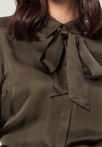 Chemise en satin vert olive avec un col et un détail de nœud papillon au niveau du cou. Présente une poche avant et une texture lisse.