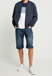 Marineblauer Bomberjacke, weißes T-Shirt mit blauer Grafik, Jeansshorts und weiße Slipper. Lässiges Outfit mit schlichtem Design und passender Passform.