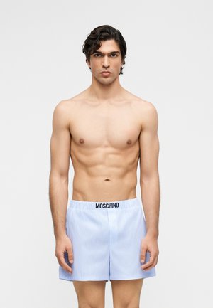 Homme torse nu aux cheveux courts et bouclés portant un boxer rayé bleu clair Moschino, debout devant un fond blanc uni.