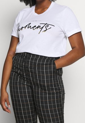 Persona con camisa blanca de manga corta con texto "moments" y pantalones negros de tiro alto con patrón de cuadros blancos y marrones, una mano en el bolsillo.
