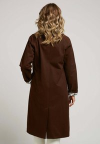 Manteau long brun avec une coupe droite, présentant un motif écossais prêt à l'emploi aux poignets et une fente au dos pour faciliter les mouvements.