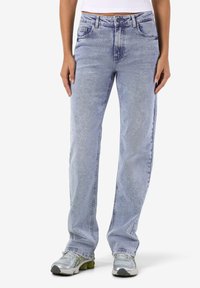 Ανοιχτό μπλε τζιν παντελόνι με ίσια γραμμή, σχεδίαση πέντε τσεπών, ξεθωριασμένη εμφάνιση και ελαφρώς φθαρμένη υφή. Φοριέται με γκρι αθλητικά παπούτσια.