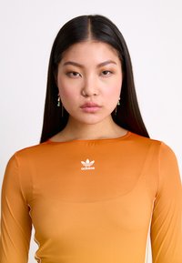 Oranžový dlouhý rukáv s hladkou texturou, s kulatým výstřihem a bílým logem Adidas na prsou.