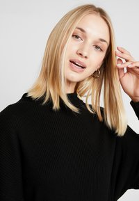 Zwarte geribde trui met een hoge halslijn, voorzien van brede mouwen en een gestructureerde afwerking, gedragen met gouden creolen.