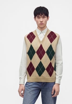 Junger Mann, der eine beige Argyle-Strickweste mit burgunderroten und grünen Rauten über einem weißen Hemd mit Knöpfen und blauen Jeans trägt.