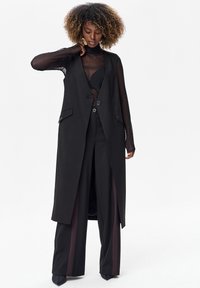 Vrouw met krullend haar die een doorschijnend zwart topje draagt, een lange mouwloze zwarte vest, zwarte broek met wijde pijpen en zijstrepen, en zwarte puntige schoenen.