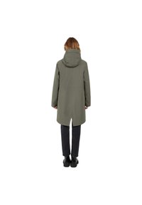 Abrigo largo verde oliva con capucha, de corte recto, con aberturas laterales y una tela suave y texturizada. Llevado sobre pantalones oscuros y botas.