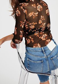 Haut black transparent à manches longues avec imprimé floral orange, porté avec un short en denim taille haute. Chaise transparente visible en arrière-plan.