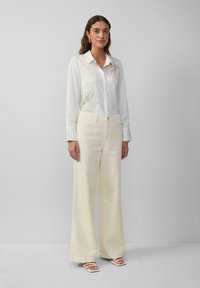 Femme portant une blouse blanche à broderies, un pantalon large crème et des sandales blanches à bout carré, adossée à un mur uni.