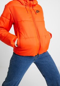 Orange puffjacka med huva, svart Nike-logotyp på bröstet och dragkedja framtill, med elastiska ärmslut och sidofickor.