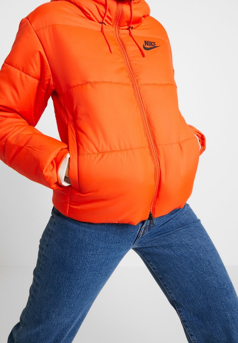 Orange puffjacka med huva, svart Nike-logotyp på bröstet och dragkedja framtill, med elastiska ärmslut och sidofickor.