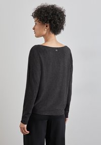 Schwarzer Langarmpullover mit weitem Ausschnitt, strukturiertem Stoff und einem kleinen Logodetail auf der Rückseite. Getragen mit schwarzen Hosen.