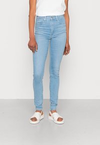 Jeans ajustados de color azul claro fabricados en denim, con una cintura de altura media, bolsillos frontales y una clásica cremallera. Combinados con sandalias blancas.