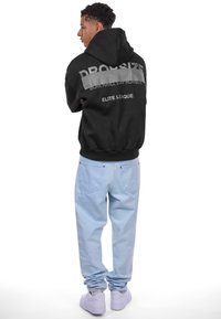 Haut à capuche noir avec "DROPSIZE" et slogans en blanc dans le dos, associé à un jean bleu clair et des baskets blanches. Vue arrière.