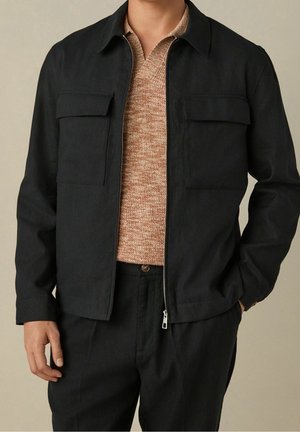 Man wearing black zip-up jacket with two chest pockets over a beige knit sweater and black pants, standing with one hand in pocket.