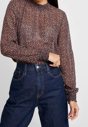 Kobieta nosząca prześwitującą, kwiecistą bluzkę z długimi rękawami, włożoną do ciemnoniebieskich, wysokich spodni jeansowych, poprawiająca mankiet na lewym nadgarstku.