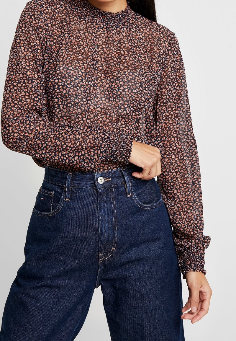 Femme portant un chemisier fleuri transparent à manches longues rentré dans un jean taille haute bleu foncé, ajustant le poignet gauche.