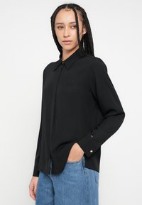 Fekete hosszú ujjú ing galléros nyakkal, aranygombos részlettel az elején és finom logóhímzéssel, világoskék farmernadrággal viselve.