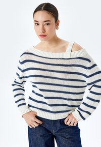 Sweter w paski w kolorze kremowym i granatowym, z szerokim, odsłoniętym dekoltem, prążkowanymi mankietami i luźnym splotem. Połączony z dżinsami.