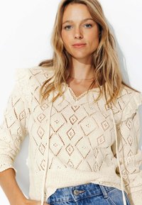 Pull en maille de couleur crème avec un motif en diamant, des accents en dentelle et un col en V. Présente des épaules froncées et un détail à cordon de serrage.