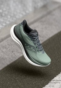 Lysegrønn og grå Saucony løpesko med hvit såle, avbildet i luften over teksturert fortau.