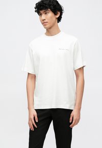 Bela bombažna majica z ohlapnim krojem, kratkimi rokavi in majhnim črnim napisom na prsih. Kombinirana s črnimi hlačami.