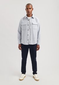 Jasnoszara koszula na guziki z dwoma kieszeniami na piersi, zestawiona z granatowymi, dopasowanymi spodniami oraz białymi sneakersami z beżowymi dodatkami. Prosty design.
