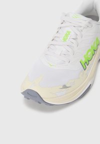 Baskets de course blanches avec une tige en mesh texturé, logo vert et accents beiges. Présentent une semelle amortissante et un design à lacets.