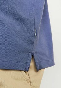 Krupan plan plavog teksturiranog polo majice na rubu s bočnim prorezom i malom etiketom marke "Jacksons", nosi se preko bež hlača.