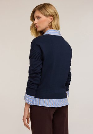 Marineblauwe trui met geribbelde details, gedragen over een lichtblauw gestreept shirt. De manchetten van het shirt zijn zichtbaar onder de zoom van de trui.