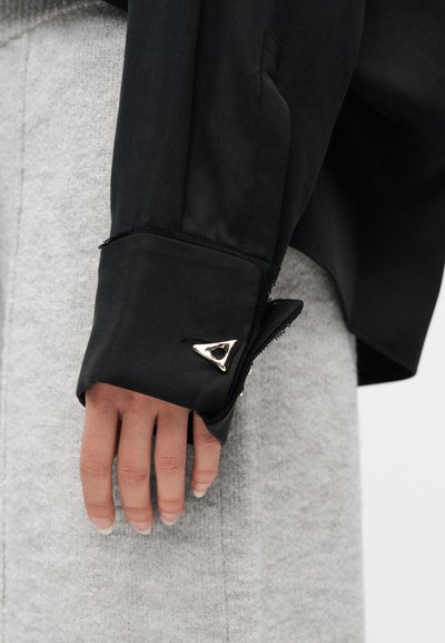 Χέρι με φυσικά νύχια δίπλα σε γκρι ύφασμα, φοράει μαύρο μανίκι με διπλωμένο μανσέτο που ασφαλίζει με ένα τριγωνικό ασημένιο κουμπί μανσετών.