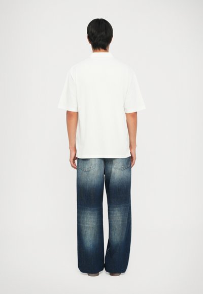 Tricou polo alb cu mânecă scurtă, asociat cu blugi din denim albastru deschis, evazați, având buzunare la spate și un efect de spălare în gradient.