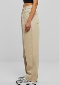 Beige široké kalhoty z bavlny s vysokým pasem, dvěma bočními kapsami a přímým střihem, v kombinaci se šedými teniskami.