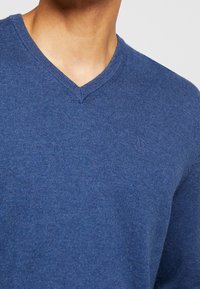 Mörkblå v-ringad tröja i mjukt stickat material, med en subtil logotyp på bröstet och ribbade ärmslut. En enkel och mångsidig design.