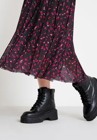 Bottes de combat noires en cuir avec une semelle épaisse, dotées de lacets et de fermetures éclair sur les côtés, associées à une jupe fluide à motif noir et rose.