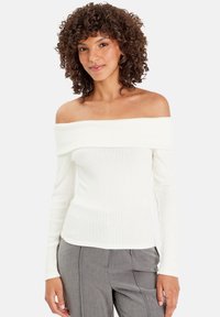 Donna con capelli ricci che indossa un maglione bianco a spalle scoperte e pantaloni grigi, in piedi davanti a uno sfondo bianco.