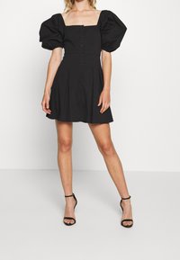 Czarna sukienka z bufkami, z kwadratowym dekoltem i zapięciem na guziki z przodu; charakteryzuje się rozkloszowanym dołem i gładką fakturą materiału.
