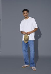 Fehér oversized póló, kék egyenes lábú farmer, bézs baseball sapkát tart a kezében. Szürke háttér; a modell magabiztosan áll, a kezei láthatók.