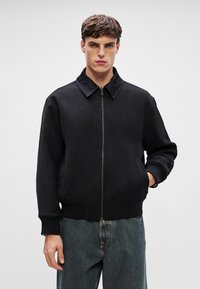 Sort bomberjakke med høj krave, frontlynlås og ribstrikkede manchetter. Stoffet ser glat ud med en mat finish. Modellen står med ansigtet fremad.