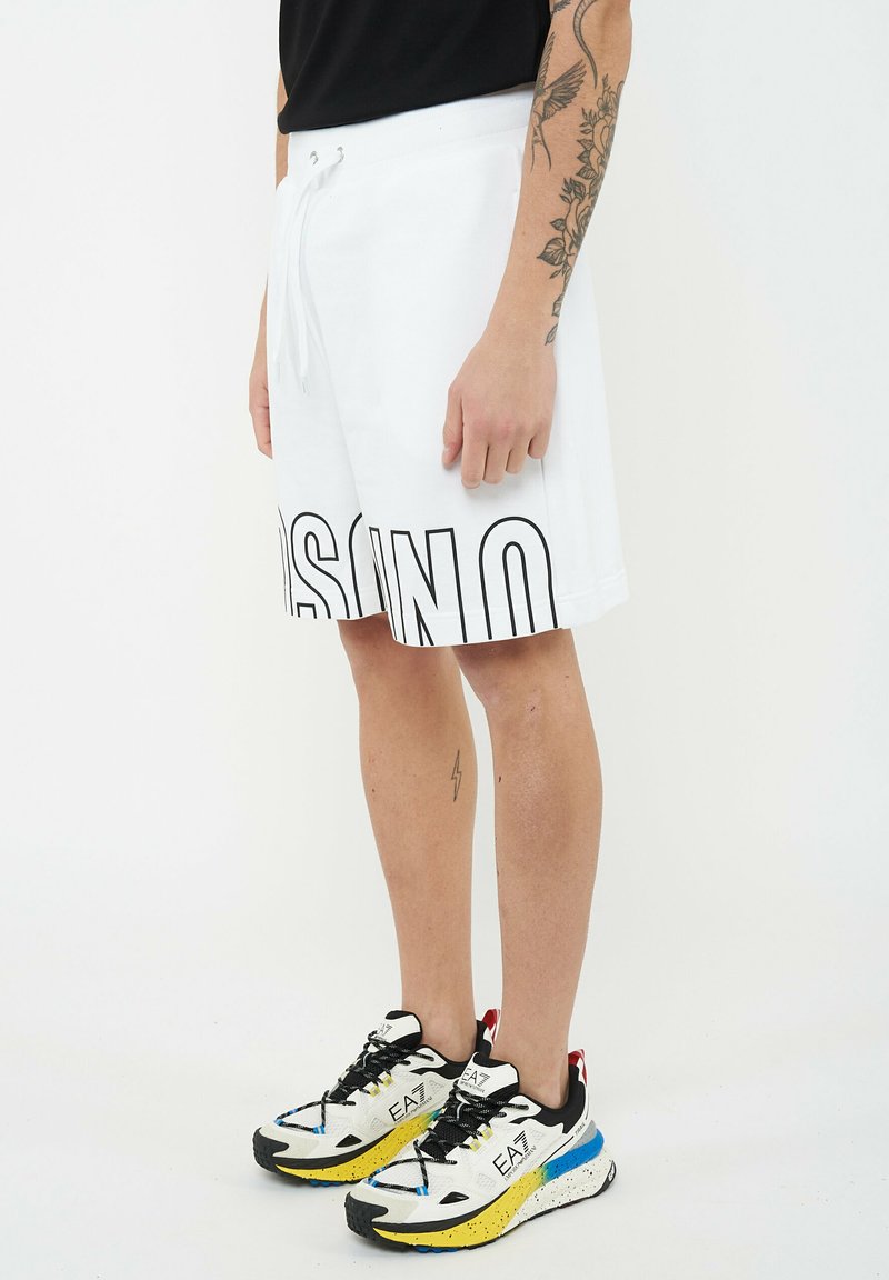 Personne portant un short blanc à cordon avec de grandes lettres noires en contour et des baskets multicolores EA7, debout devant un fond uni.